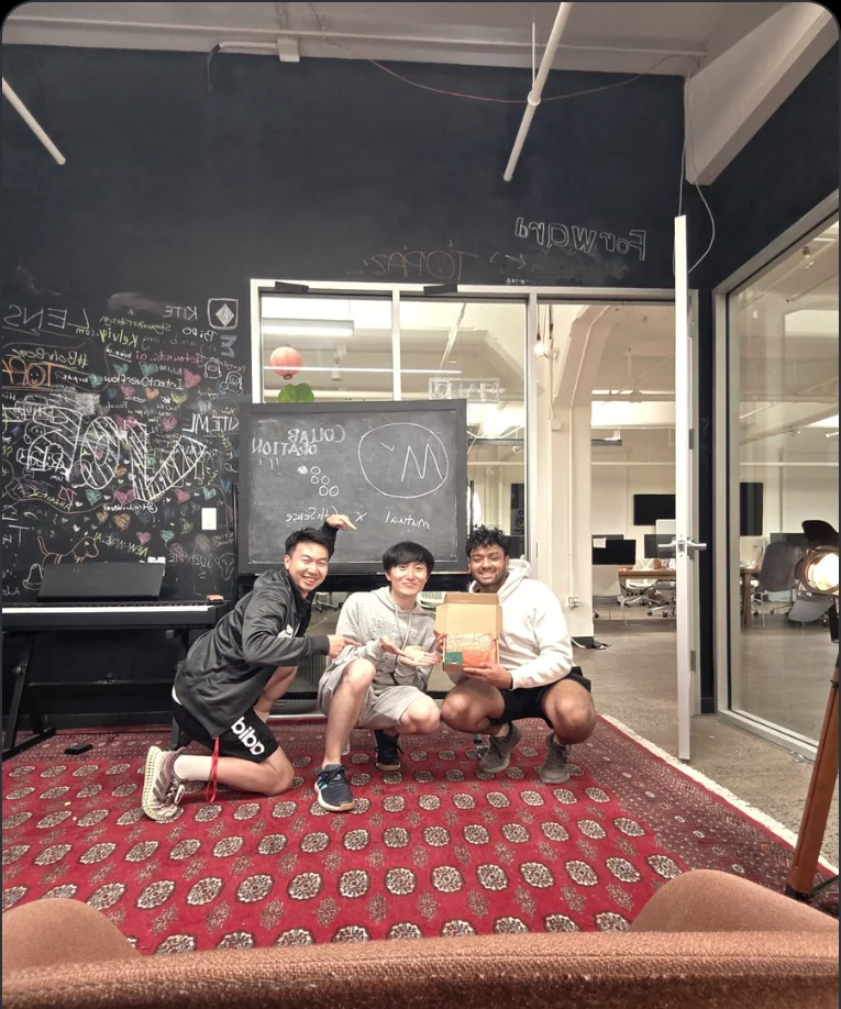 Three people crouched on a patterned rug in an office, holding a small opened box, in front of a chalkboard