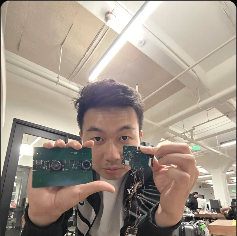 A person holding up two bare circuit boards side by side: the wider development prototype with two lens modules on it, and the smaller stereo board